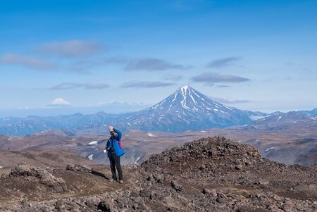 young man traveler photographer at the top of the volcano mountain shows excellent, rejoices, volcanic view in the crater of the volcano, the traveler at the height, the view of the mountain topsの写真素材