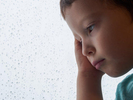 A lonely child sits by the window, looking out thoughtfully. Soft light through the glass, creating an atmosphere of comfort and melancholy.の写真素材