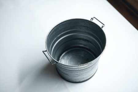 A solitary galvanized metal bucket rests on a white surface, bathed in soft light.の素材