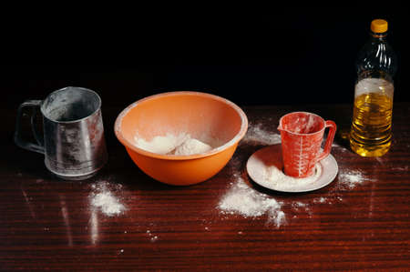 Orange cup, measuring cup, and a steel sieve, sunflower oil stand on a wooden table on a black background. Flour.の写真素材