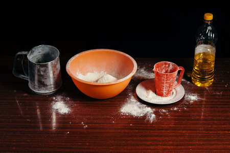 Orange cup, measuring cup, and a steel sieve, sunflower oil stand on a wooden table on a black background. Flour.の写真素材