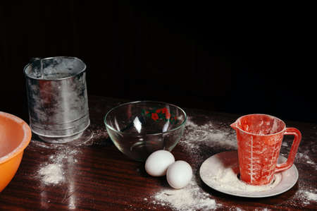 Orange cup, measuring cup, and a steel sieve, two eggs stand on a wooden table on a black background. Flour.の写真素材