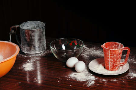 Orange cup, measuring cup, and a steel sieve, two eggs stand on a wooden table on a black background. Flour.の写真素材