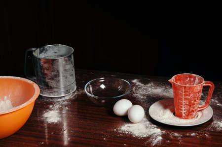Orange cup, measuring cup, and a steel sieve, two eggs stand on a wooden table on a black background. Flour.の写真素材