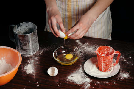 A girl in an apron in a dark kitchen breaks the egg into a bowl.の写真素材