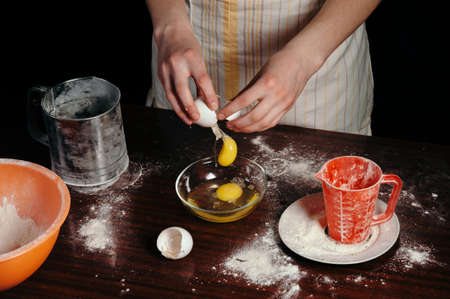 A girl in an apron in a dark kitchen breaks the egg into a bowl.の写真素材