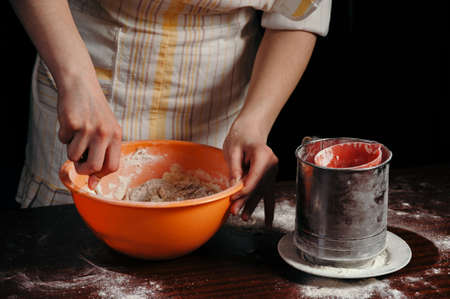 A girl in an apron in a dark kitchen kneads the dough in an orange cup.
