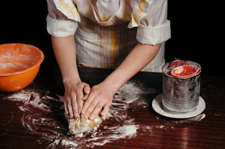 A girl in an apron in a dark kitchen kneads the dough with her hands on a wooden kitchen table. Close-up.の写真素材