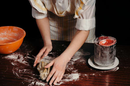 A girl in an apron in a dark kitchen kneads the dough with her hands on a wooden kitchen table. Close-up.の写真素材