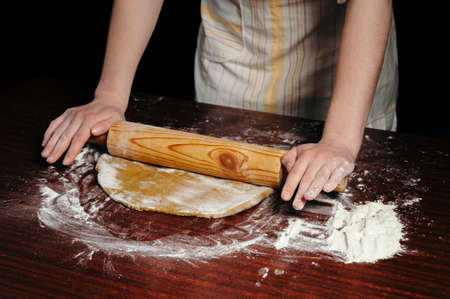 A woman in the kitchen is rolling a dough on a wooden table with a wooden rolling pin.の写真素材