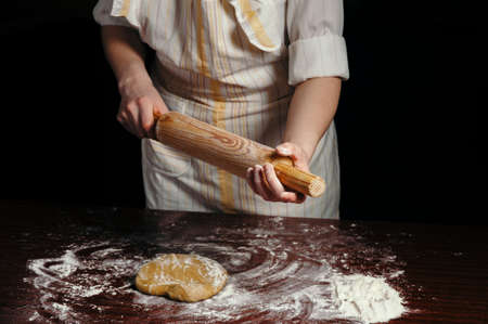 A woman in a light apron in a dark kitchen. She rubs the wooden rolling pin with flour.の写真素材