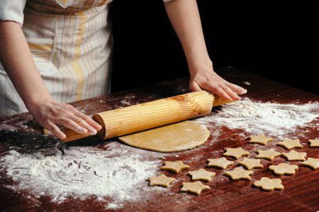 A woman in the kitchen is rolling a dough on a wooden table with a wooden rolling pin. She molds cookies in the form of asterisks.の写真素材