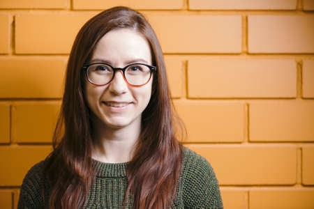 Beautiful red-haired girl wearing glasses and a green sweater is smiling against a yellow brick wall background.の写真素材