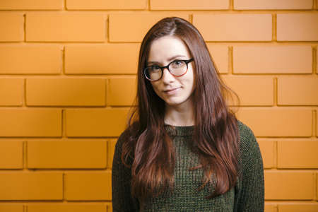 Beautiful red-haired girl wearing glasses and a green sweater is smiling against a yellow brick wall background.の写真素材