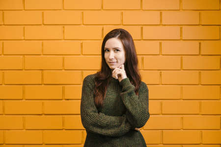 Beautiful red-haired girl wearing glasses and a green sweater is smiling against a yellow brick wall...