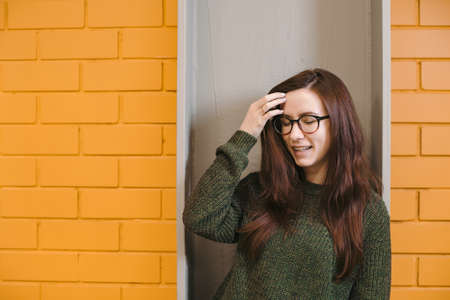 Beautiful red-haired girl wearing glasses and a green sweater is smiling against a yellow brick wall background.の写真素材