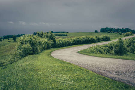 Dramatic green landscape with asphalt road in towards mountains under cloudy sky.の写真素材