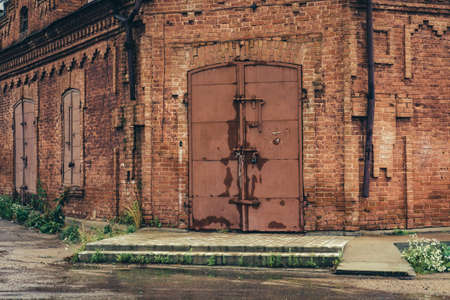 Old red brick building with huge iron locked gate in rainy weather. Green grass grows  through old wet asphalt around building.の写真素材