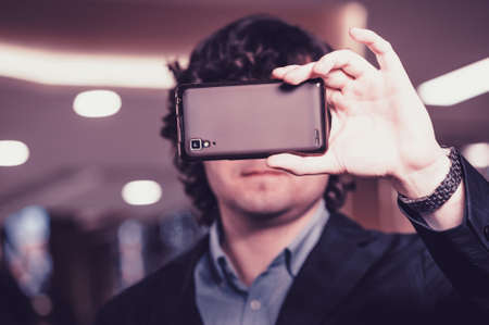 Business man looking at smartphone screen. Guy hides behind the mobile phone. Magenta tone.の写真素材