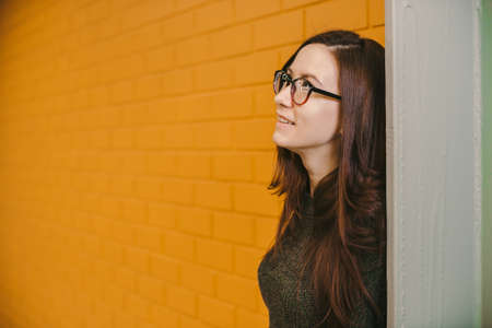 Beautiful cheerful pensive dreaming girl with long red hair dyed with henna, in green blouse and red glasses, on background of yellow brick wall, looks in sideways and awayの写真素材