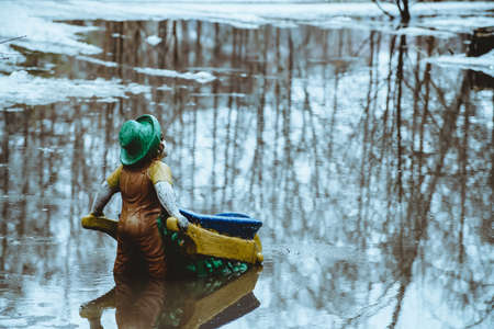 Figurine in form of gnome stands with his back with barrow in water in spring in Russian reality. Snow is melting, creating huge puddle. Atmosphere as in fairy tale.の写真素材