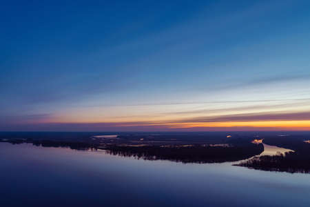 Beautiful river with big island with trees under predawn sky. Bright yellow stripe in picturesque cloudy sky. Early blue sky reflected in water. Colorful morning atmospheric image of majestic nature.の写真素材