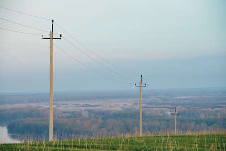 Atmospheric landscape with power lines in green field on background of river under blue sky. Electric pillars with copy space. Wires of high voltage above ground. Electricity industry.の写真素材