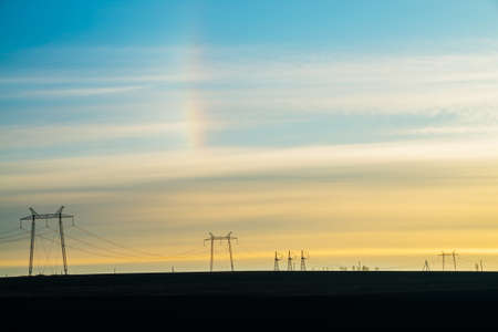 Power lines on background of sunny sky with rainbow close-up. Silhouette of electric pole with copy space. Wires of high voltage above ground. Electricity industry.の写真素材