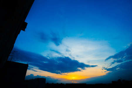Atmospheric blue cloudy sky behind silhouettes of city buildings. Cobalt and orange background of sunrise with dense clouds and bright yellow sunny light for copy space. Cyan heaven above clouds.の写真素材