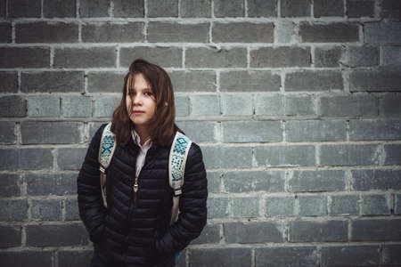 Schoolgirl with backpack stands against gray brick wall with sad emotion in cloudy weather with copy space. Atmospheric portrait of melancholic teenage girl in faded tones. Depressed young girl.の写真素材