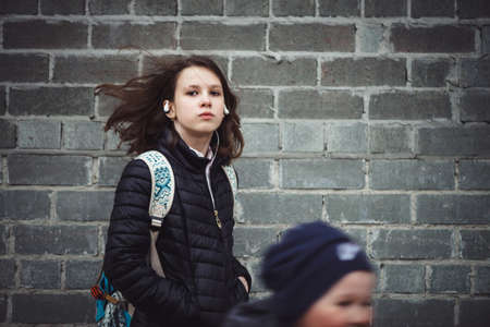Schoolgirl with backpack with flying hair and headphones against gray brick wall with copy space....