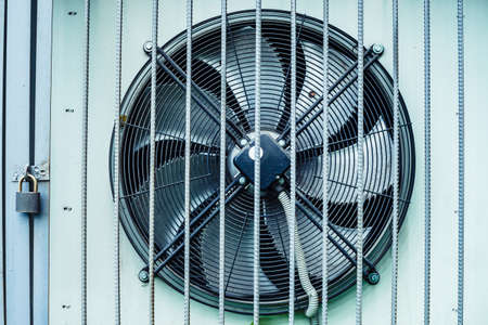 Outdoor industrial air conditioner cooler behind fence is locked on padlock with copy space. Large rotating cooling fan close-up. Conceptual background of industry equipment.の写真素材