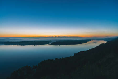 View from high shore on broad river. Riverbank with forest under thick fog. Dawn reflected in water. Yellow glow in picturesque predawn sky. Colorful morning atmospheric landscape of majestic nature.の写真素材
