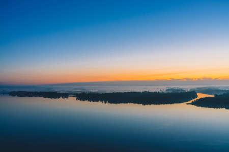 Broad river flows along diagonal shore with forest under thick fog. Early blue sky reflected in water. Yellow glow in picturesque predawn sky. Colorful morning atmospheric landscape of majestic natureの写真素材