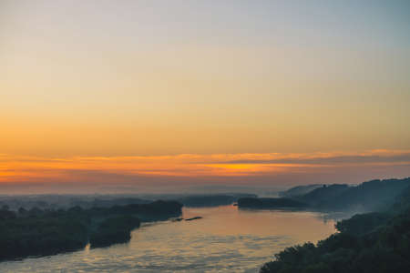 View from high shore on river. Riverbank with forest under thick fog. Gold dawn reflected in water. Orange glow in picturesque predawn sky. Mystical morning atmospheric landscape of majestic nature.の写真素材