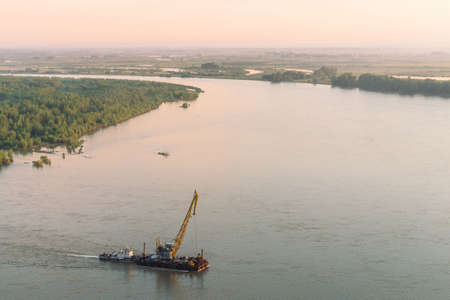 Beautiful river landscape with green shores and ship with copy space. Towboat tows barge with crane along riverbank. Dawn reflexed on calm glare water surface. Pink sunset sky on background.の写真素材