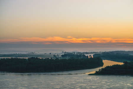 Early blue sky reflected in river water. Riverbank with forest under predawn sky. Orange stripe in picturesque sky. Fog hid trees on island. Colorful morning atmospheric landscape of majestic nature.の写真素材