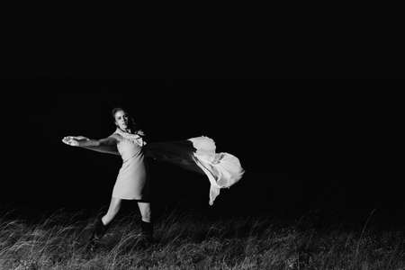 Amazing girl dancing in dress with big white shawl in darkness in grayscale. Beautiful female dancer with good mood in night. Unimaginable mystic dance on nature in dark in monochrome.の写真素材