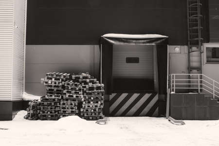 Cargo lift for storage with white black diagonal lines warning sign in grayscale. Wooden pallets on snow. Door for loading goods with copy space. Wall of warehouse in winter. Monochrome building.の写真素材