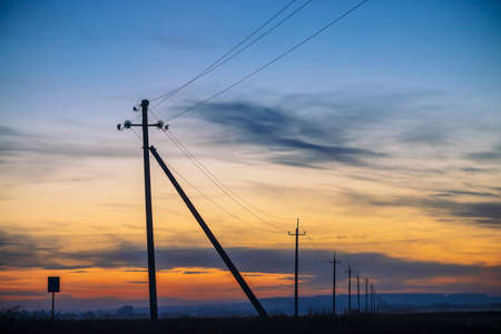 Power lines in field on sunrise background. Silhouettes of poles with wires at dawn. Cables of high...