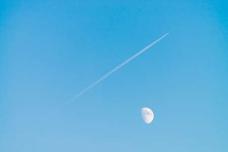 Condensation track of jet above moon in clear blue day sky. Minimalist blue background. Plane flies up diagonally. Airplane is flying in air space. White moon in cyan sky.の写真素材