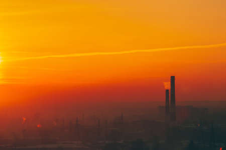 Smog among silhouettes of buildings on sunrise. Smokestack in dawn sky. Environmental pollution on sunset. Harmful fumes from stack above city. Mist urban background with warm orange yellow sky.の写真素材