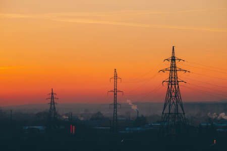 Power lines in city on dawn. Silhouettes of urban buildings among smog on sunrise. Cables of high voltage on warm orange yellow sky. Power industry at sunset. City power supply. Mist urban background.の写真素材