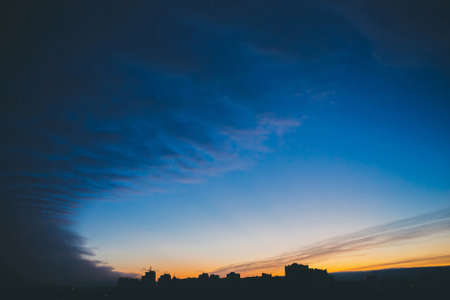 Cityscape with wonderful varicolored vivid dawn. Amazing dramatic blue cloud sky above dark silhouettes of city building roofs. Atmospheric background of orange sunrise in overcast weather. Copy spaceの写真素材