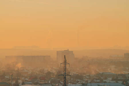Power lines in city on dawn. Silhouettes of urban buildings among smog on sunrise. Cables of high voltage on warm orange yellow sky. Power industry at sunset. City power supply. Mist urban background.の写真素材