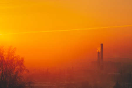 Smog among silhouettes of buildings on sunrise. Smokestack in dawn sky. Environmental pollution on sunset. Harmful fumes from stack above city. Mist urban background with warm orange yellow sky.の写真素材