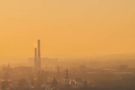 Smog among silhouettes of buildings on sunrise. Smokestack in dawn sky. Environmental pollution on sunset. Harmful fumes from stack above city. Mist urban background with warm orange yellow sky.の写真素材