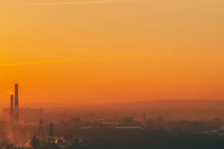 Smog among silhouettes of buildings on sunrise. Smokestack in dawn sky. Environmental pollution on sunset. Harmful fumes from stack above city. Mist urban background with warm orange yellow sky.の写真素材