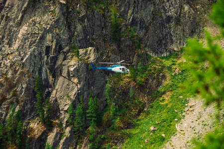 Helicopter in air against background of giant green rock covered with vegetation in tropical. Rescue mission in mountains. Aerial tourism in warm climate. Trees and grass on mountainside.の写真素材