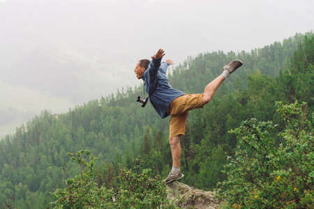 Mad tourist on mountain peak. Joyful traveler stands on one leg over abyss. Crazy man is jumping on rocky summit. Positive person in flight over precipice. Exciting rainy moment. Freedom in highlands.の写真素材
